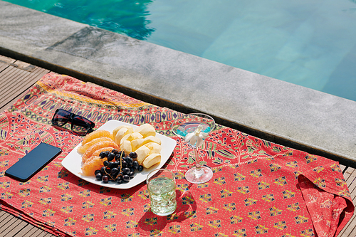 Glasses with refreshing cocktails and plate with fruits on blanket nest to swimming pool