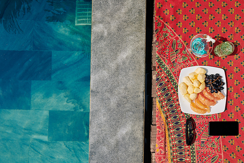 Plate with fruits, delicious cocktails, smartphoe and sunglasses on side of pool with turquoise water, view from above
