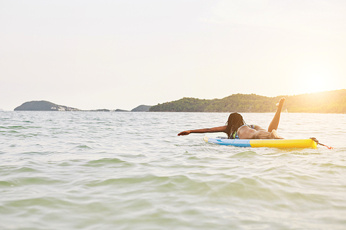 Black young woman swimming on sup board in the ocean in rayS of declining sun