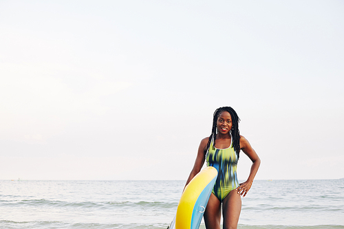 Portrait of happy smiling young fit Black woman carrying sup board when walking out of ocean