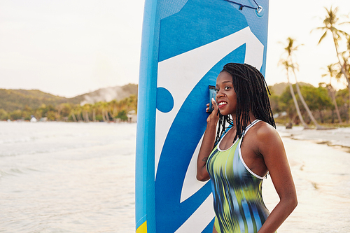 Happy pretty young Black fit woman standing on beach with sup board and looking at sea