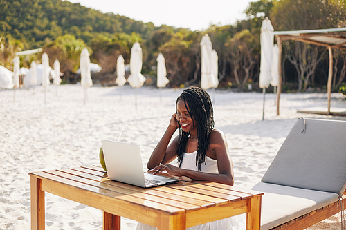 Smiling pretty female freelancer sitting at desk on sandy beach and working on laptop