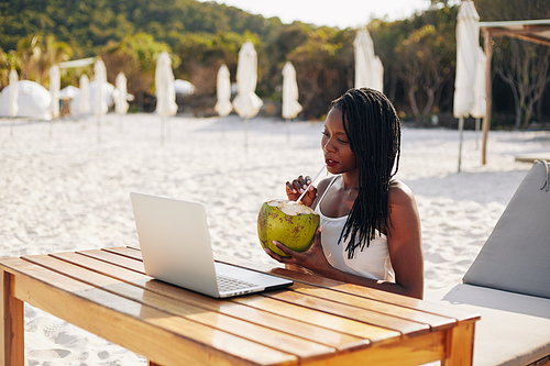 Pretty young Black woman with dreadlocks drinking coconut cocktail and watching webinar on laptop when sitting at table outdoors