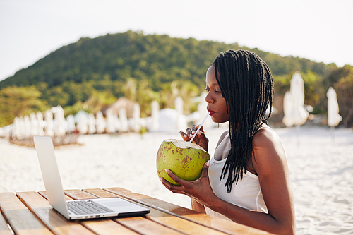 Beautiful young Black woman drinking refreshing coconut water when sitting on beach and reading article on laptop screen