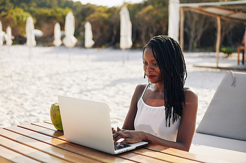 Pretty young woman sitting at wooden table on sea beach and working on laptop