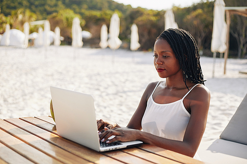 smiling pretty young travel vlogger sitting at wooden table on beach and working on laptop when making post for her Vlog