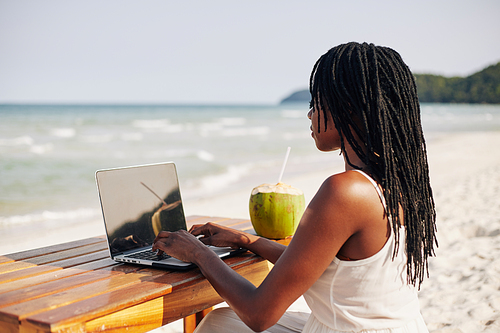 Black young woman with dreadlocks working on computer when sitting at desk on the beach