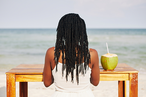 Young Black woman with dreadlocks sitting at table with coconut water, working on laptop ang enjoying sea view