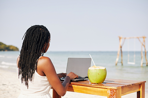 Young woman sitting at desk on sandy beach, drinking cocnut cocktail and answering e-mails on her laptop