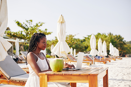 Pretty young woman sitting at table on sandy beach, enjoying fresh coconut cocktail and working on laptop