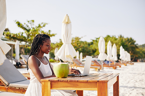 Smiling pretty young Black woman sitting at table on sandy beach with coconut cocktail and working on laptop