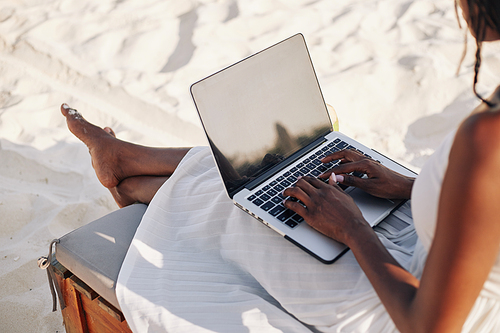 Young woman with laptop sitting on chaise-lounge on beach and working remotely