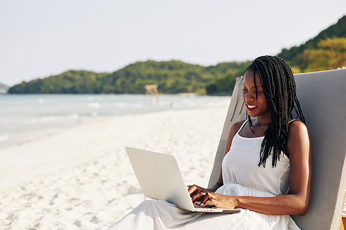 Portrait of pretty smiling young Black woman resting on chaise-lounge on sandy beach and working on laptop