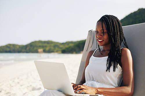 Relaxed beautiful young Black woman resting on beach on sunny day and working on laptop