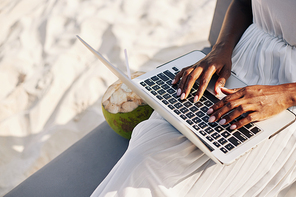 Close-up image of woman sitting on sandy beach and working on laptop, coding or answering e-mails
