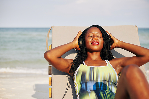 Happy beautiful young Black woman relaxing in chaise-lounge and listening to music in headphones