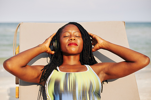 Beautiful young Black woman closing eyes and putting on headphones when sunbathing on chaise-lounge on beach