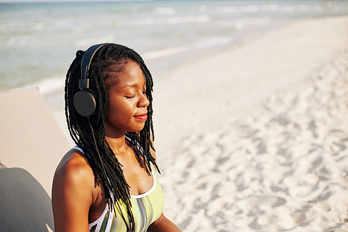 Smiling yuong woman listening to relaxing music in headphones when meditating on beach