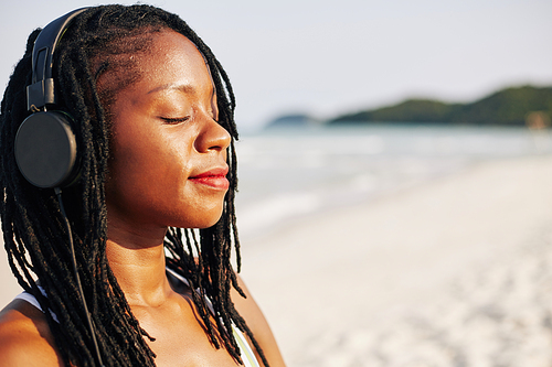 Face of beautiful young Black woman smiling and closing eyes when listening to relaxing music in her headphones and enjoying sea breeze