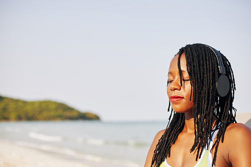 Pretty young Black woman in headphones standing on beach, relaxing and breathing fresh air