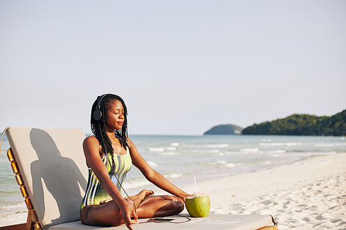 Young woman sitting on chaise-lounge in lotus position and listening to relaxing music in headphones