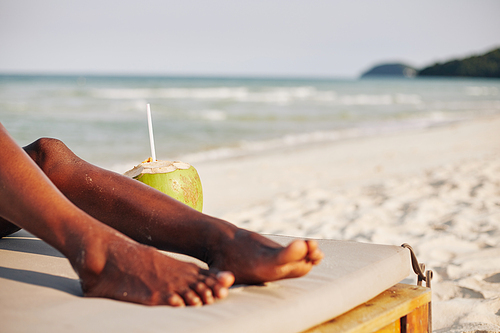 Cropped image of young Black woman resting on chaise-lounge and drinking refreshing coconut water on sea beach
