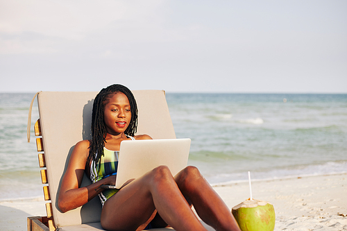 Positive attractive young Black woman resting on chaise-lounge and watching tv series on laptop screen