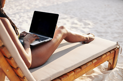 Young woman sitting on chaise-lounge on sunny sandy beach and working on laptop