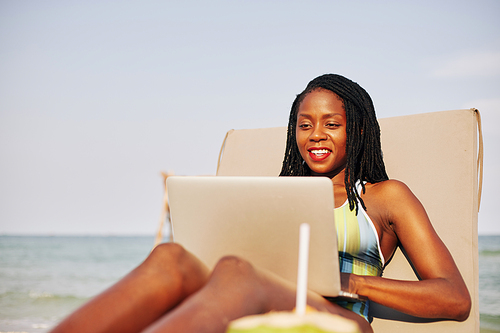 Happy pretty young Black woman sitting on chaise-lounge and answering e-mails of coworkers