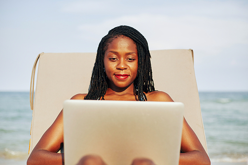Portrait of young pretty Black woman relaxing on chaise-lounge and working on laptop on beach