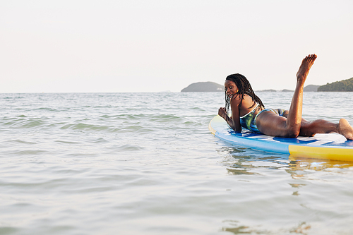 Beautiful young Black woman in swimming suit relaxing on floating sup board on sea waves and turning back to camera