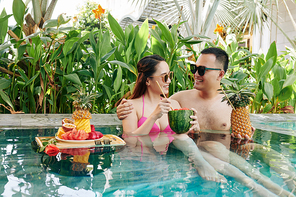Happy young Asian couple spending honeymoon in spa hotel with swimming pool and drinking cocktails
