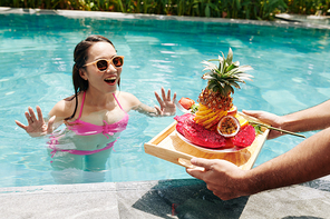 Young man bringing tray with delicious breakfast and rose flower to his excited girlfrined swimming in pool