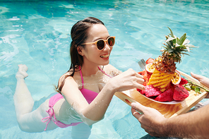 Waiter bringing tray with fresh fruits to smiling pretty young woman swimming in pool