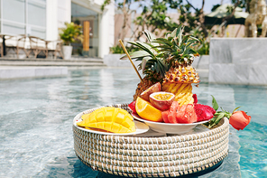 Plates with tasty sweet fruits on wicker tray floating in swimming pool of hotel