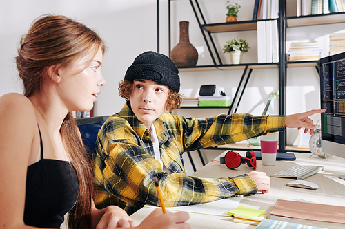 Teenage boy in plaid shirt and hat pointing at computer screen with programming code when doing homework with his sister or friend at home