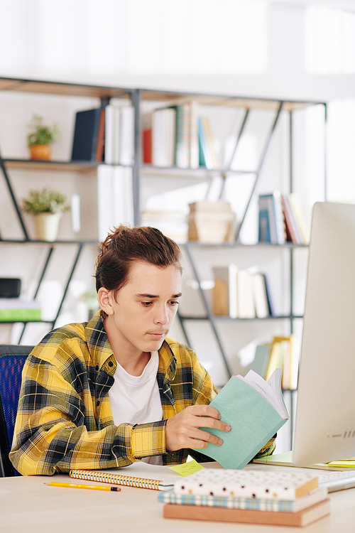 Serious teenage boy in black and yellow shirt sitting at desk at home and reading students books for upcoming classes