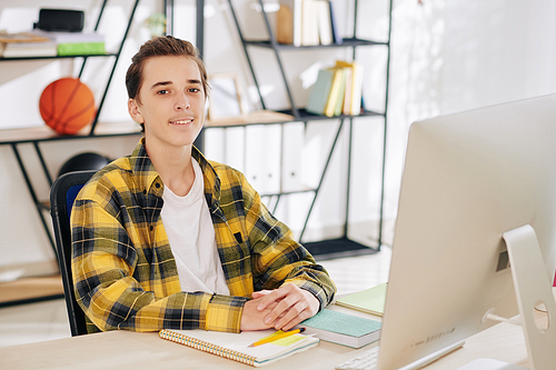 Portrait of smiling teenage boy sitting at his table in front of computer with books and textbooks ready for online lesson