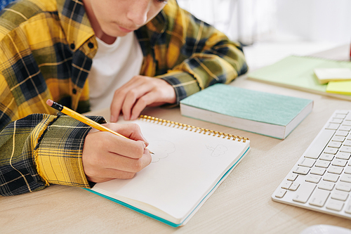 Concentrated teenage boy drawing in notebook when watching online class at home