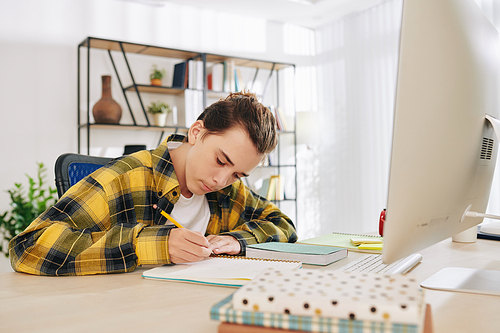 Serious concentrated teenger drawing or writing in notebook when sitting at his desk at home