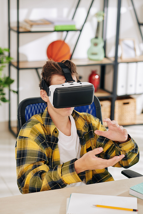 Teenage boy sitting at his table and testing new application in virtual reality headset