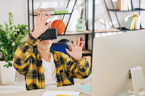 Teenage boy sitting at his desk in front of computer and testing application for VR glasses he created