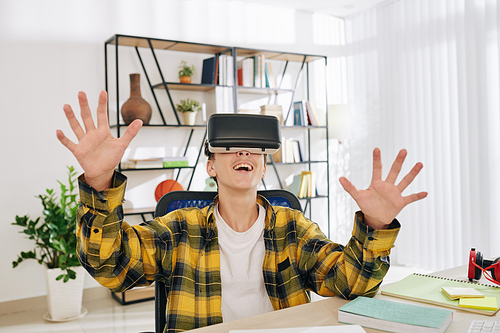 Excited happy teenage boy enjoying playing game in virtual reality headset when sitting at his tablet