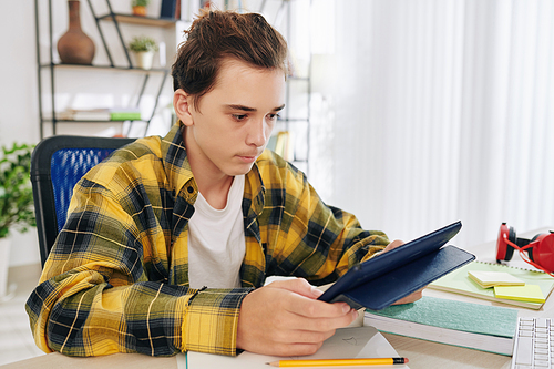 Serious teenage boy reading students book on tablet computer and homeschooling due to coronavirus pandemic