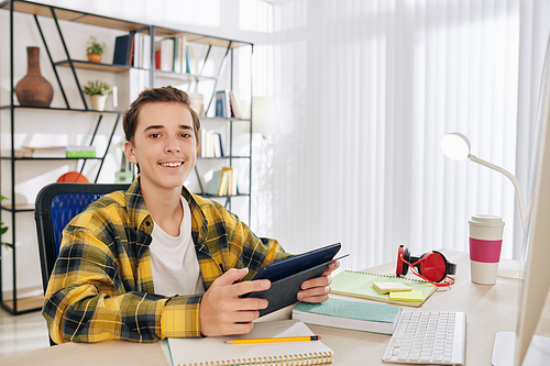 Portrait of cheerful teenage boy sitting at his desk with digital tablet in hands and smiling at camera