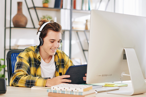 Excited teenage boy in headphones watching tv series on tablet computer instead of doing homework on computer