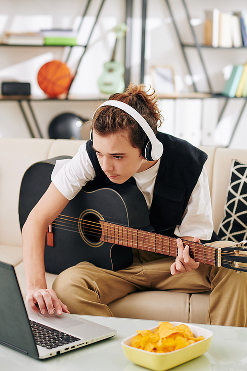 Teenage boy in wireless headphones eating potato chips when learning how to play new song on acoustic guitar