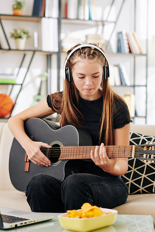Talented creative teenage girl in headphones sitting on sofa in front of opened laptop and playing guitar