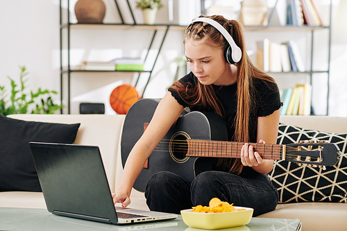 Creative serious teenage girl in wireless headphones playing guitar and writing new song on laptop