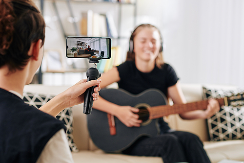 Teenage boy using smartphone on monopod when filming his sister or friend playing guitar and singing song she wrote